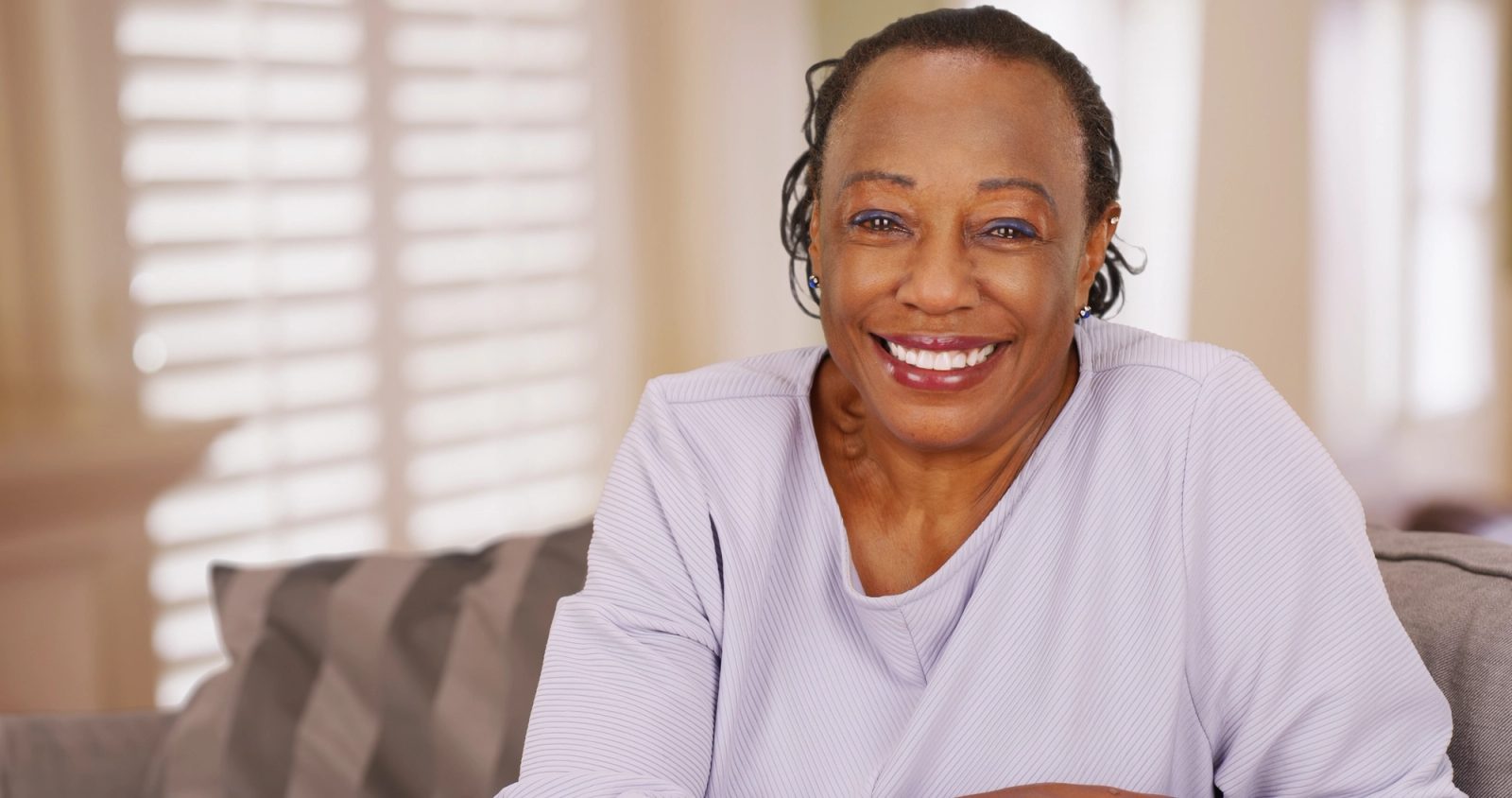 A smiling older woman with braided hair sits on a couch