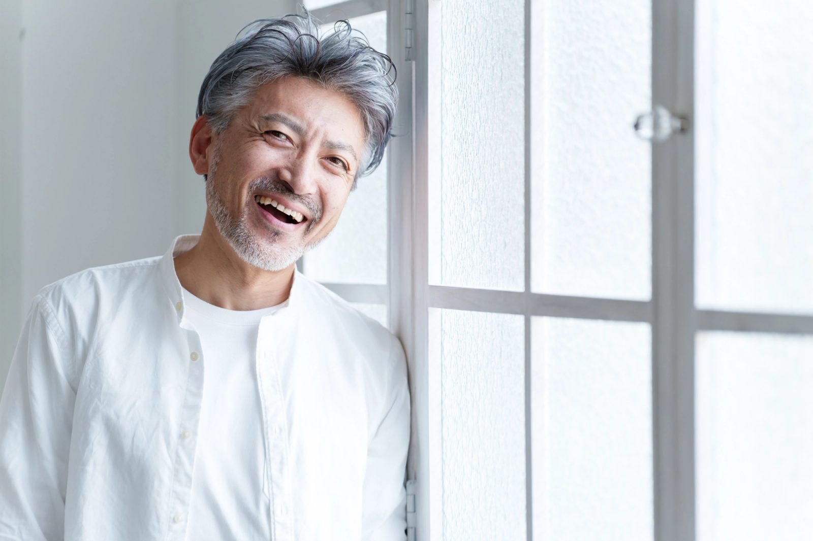 A smiling man with gray hair and a beard leans against a window